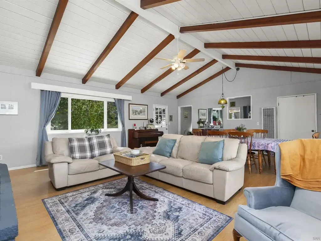Living room with tall ceiling, wood beams, couch, chair, coffee table, and dining area.
