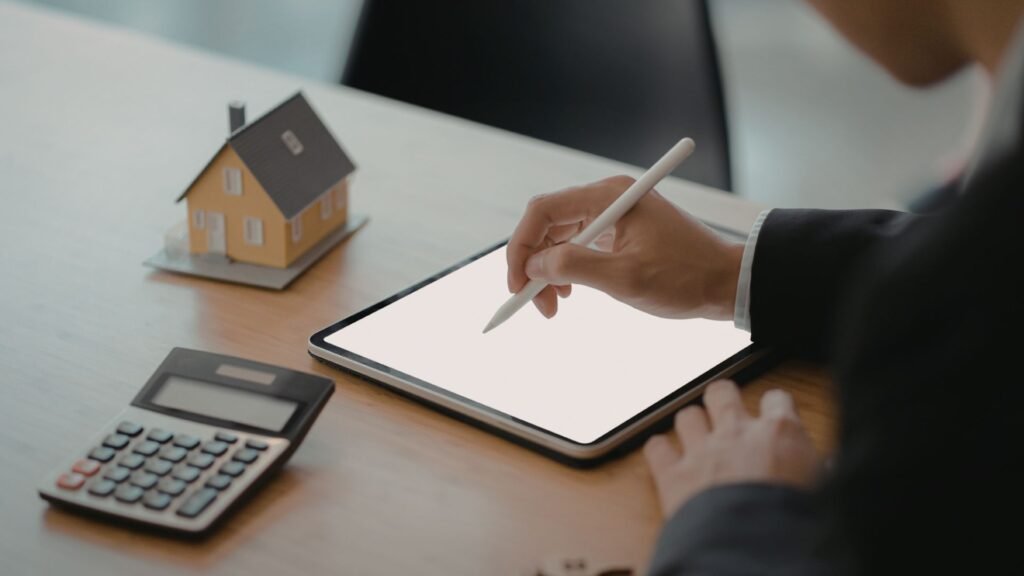 Person writing on a tablet with a stylus beside a calculator and a small house model.